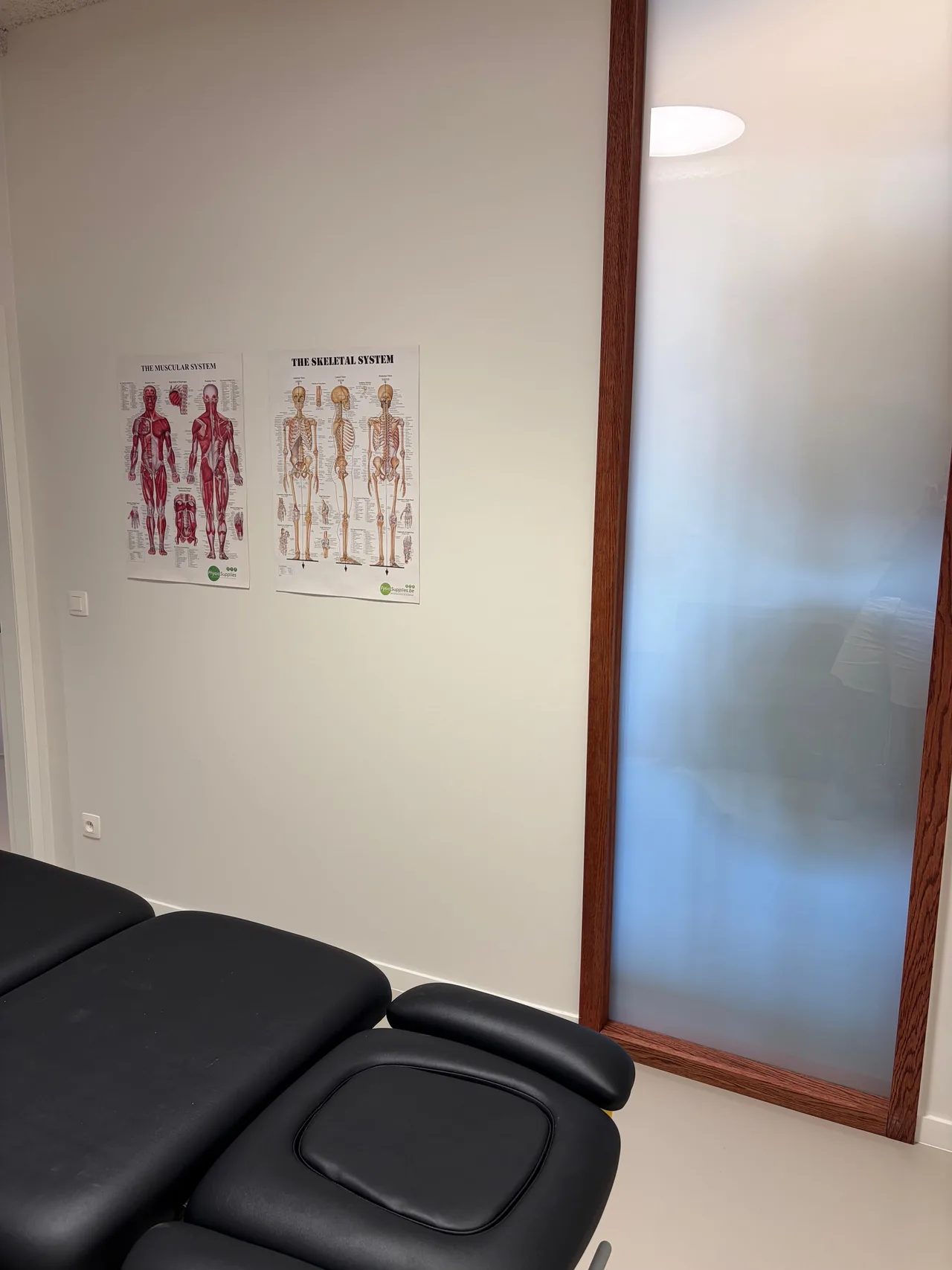 Behandelruimte met een zwarte behandeltafel en anatomische posters aan de muur